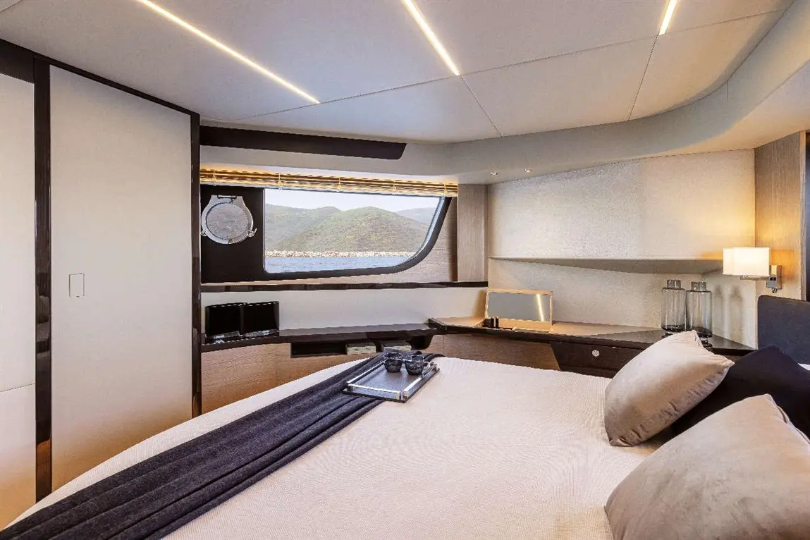 Modern yacht bedroom with a large window view, neatly made bed, and sleek furniture, illuminated by recessed lighting.